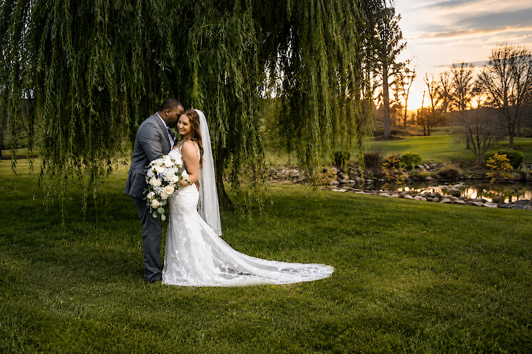Bride and groom standing under a large tree, holding each other, with a sunset and pond in the background.