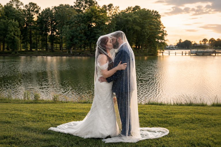 Couple in wedding attire under a veil, standing close by a lake at sunset, embracing each other.