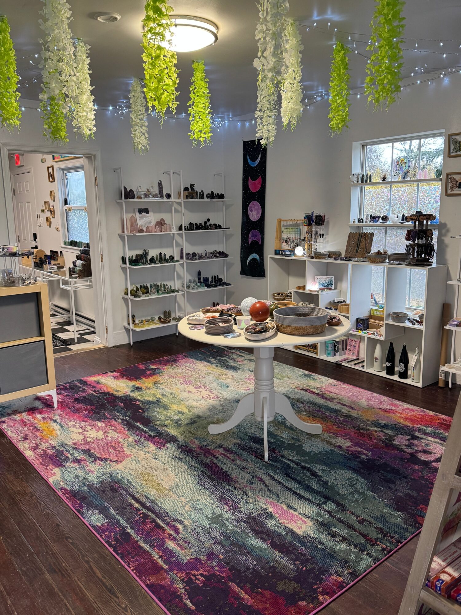 Living room with colorful rug, white shelves, and a table with food and drinks, decorated with hanging green plants.