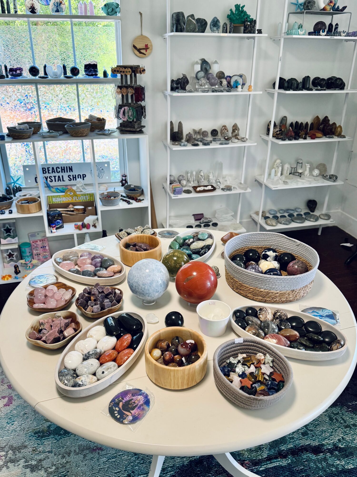 Table with various bowls of polished stones and minerals, shelves with more stones and collectibles in background.