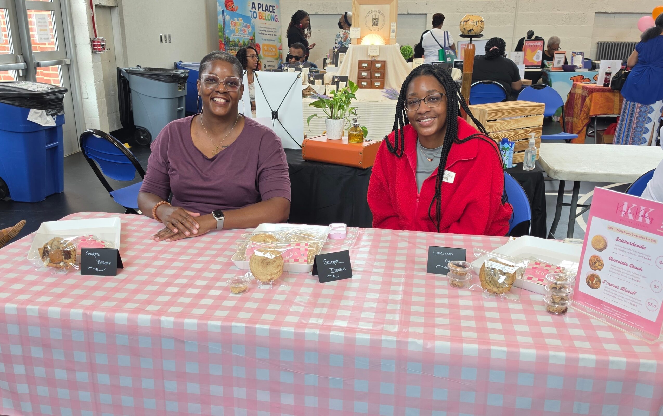 Two women sit behind a table with a pink checkered tablecloth at an indoor event, smiling at the camera.