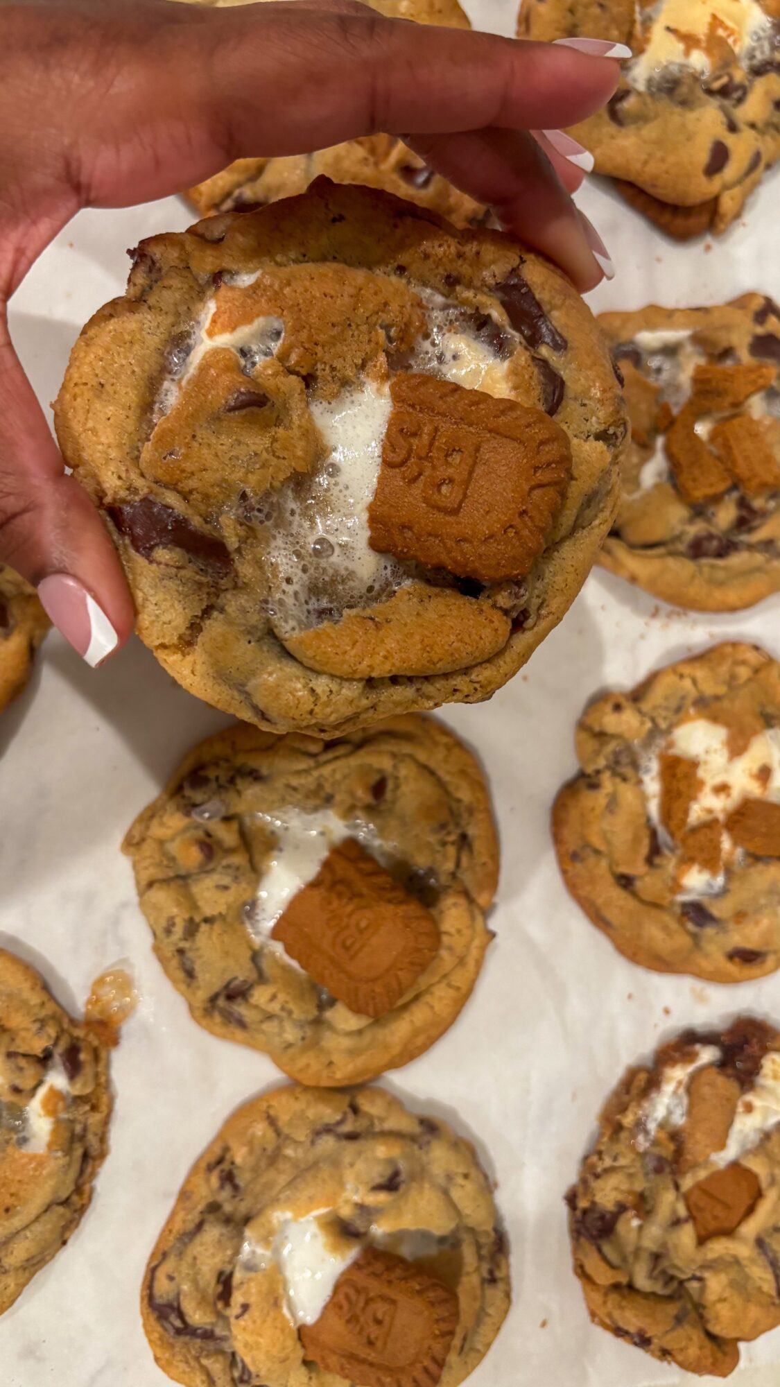 Hand holding a cookie topped with a cookie piece, surrounded by similar cookies on parchment paper.