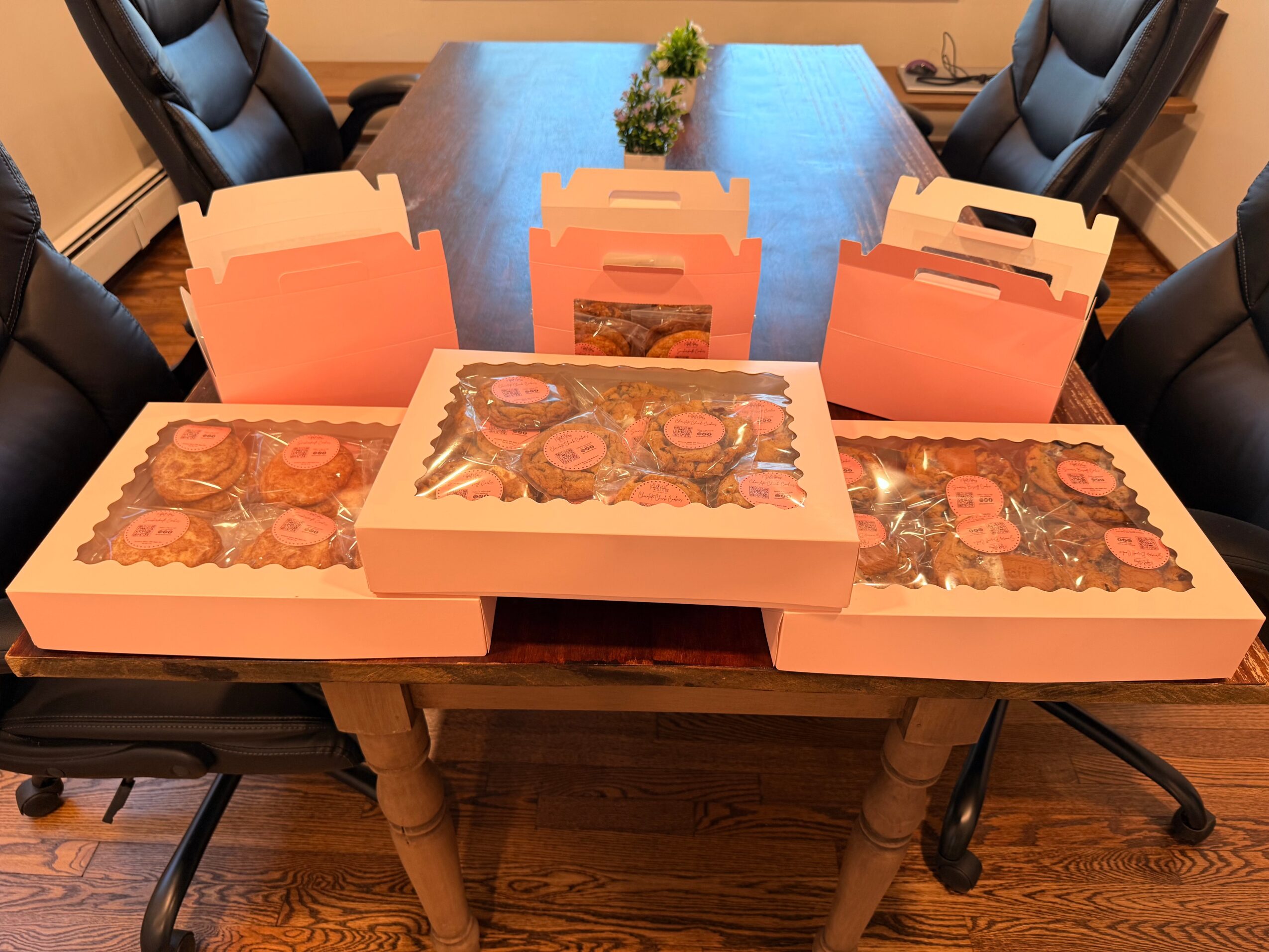 Table with pink boxes and trays of baked goods, surrounded by black chairs in a conference room.