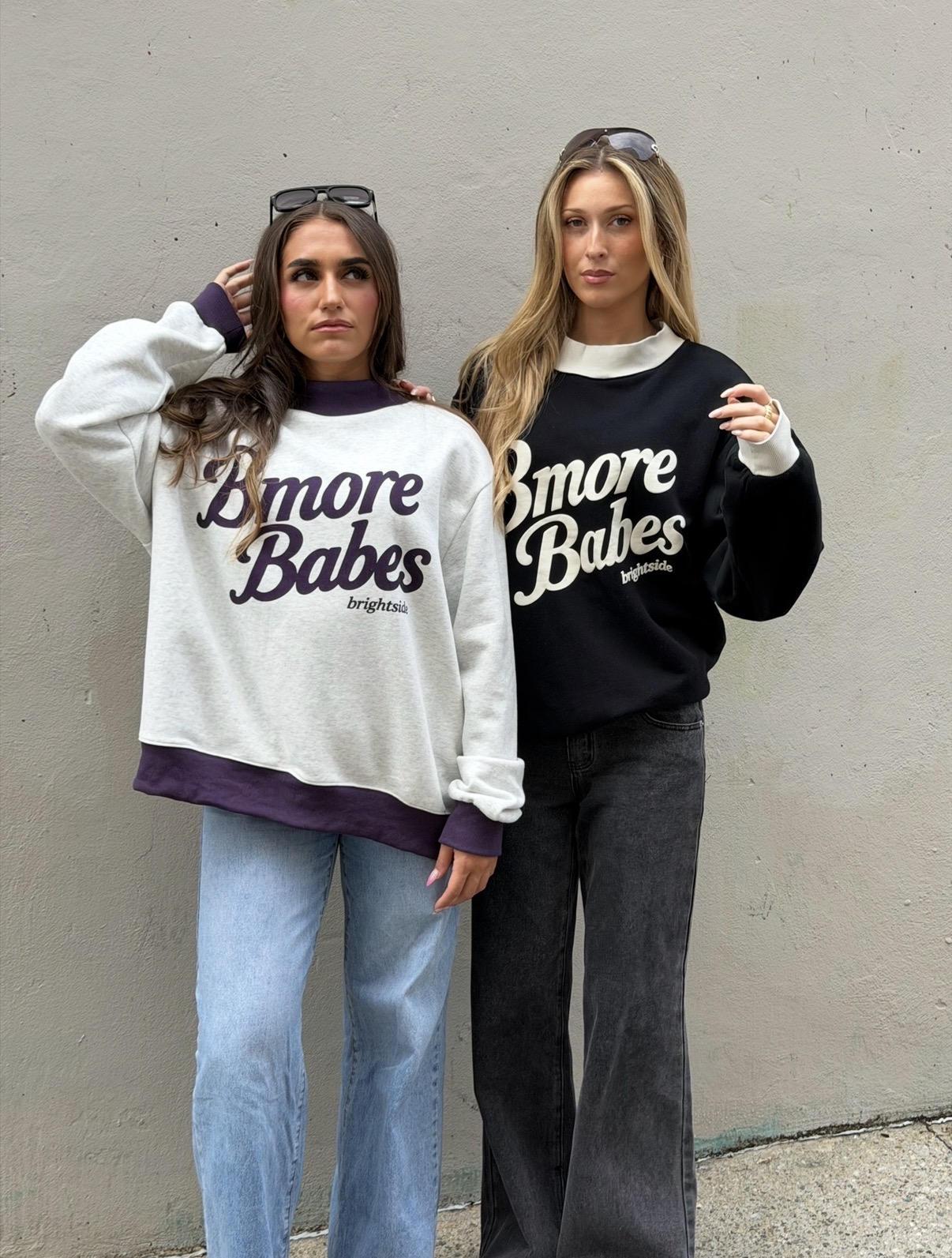Two women stand against a plain wall, wearing matching sweatshirts with 'Bmore Babes' text, one in white, the other in black.