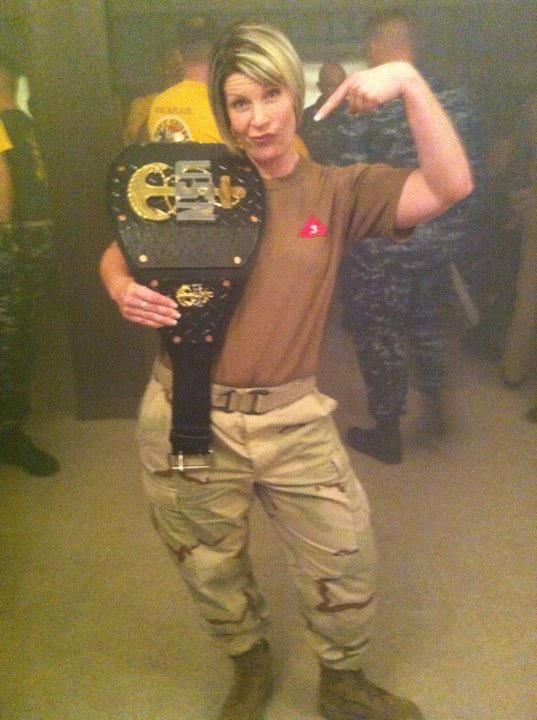 Person with short blonde hair taking a mirror selfie, flexing arm, holding a championship belt, in a room with other people.