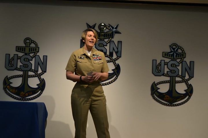 Woman in military uniform standing and smiling on stage with USN logos behind her.