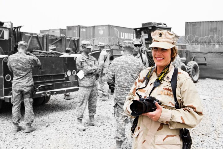 Woman in military uniform holding a camera, smiling, with soldiers and vehicles in background, black and white with color focus on woman.