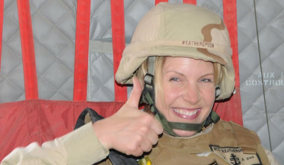Woman in military gear smiling and giving a thumbs-up, wearing a helmet and uniform, with a red and gray background.