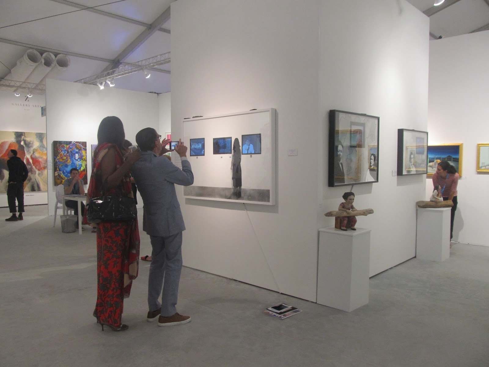 People viewing artwork in an art gallery with white walls and various paintings displayed. Two people are taking photos of art pieces.