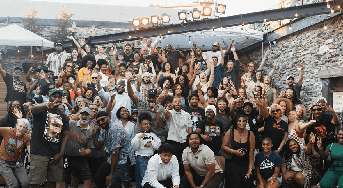 Large group of people gathered outdoors, some raising their hands, in front of a stone wall and stage lights.