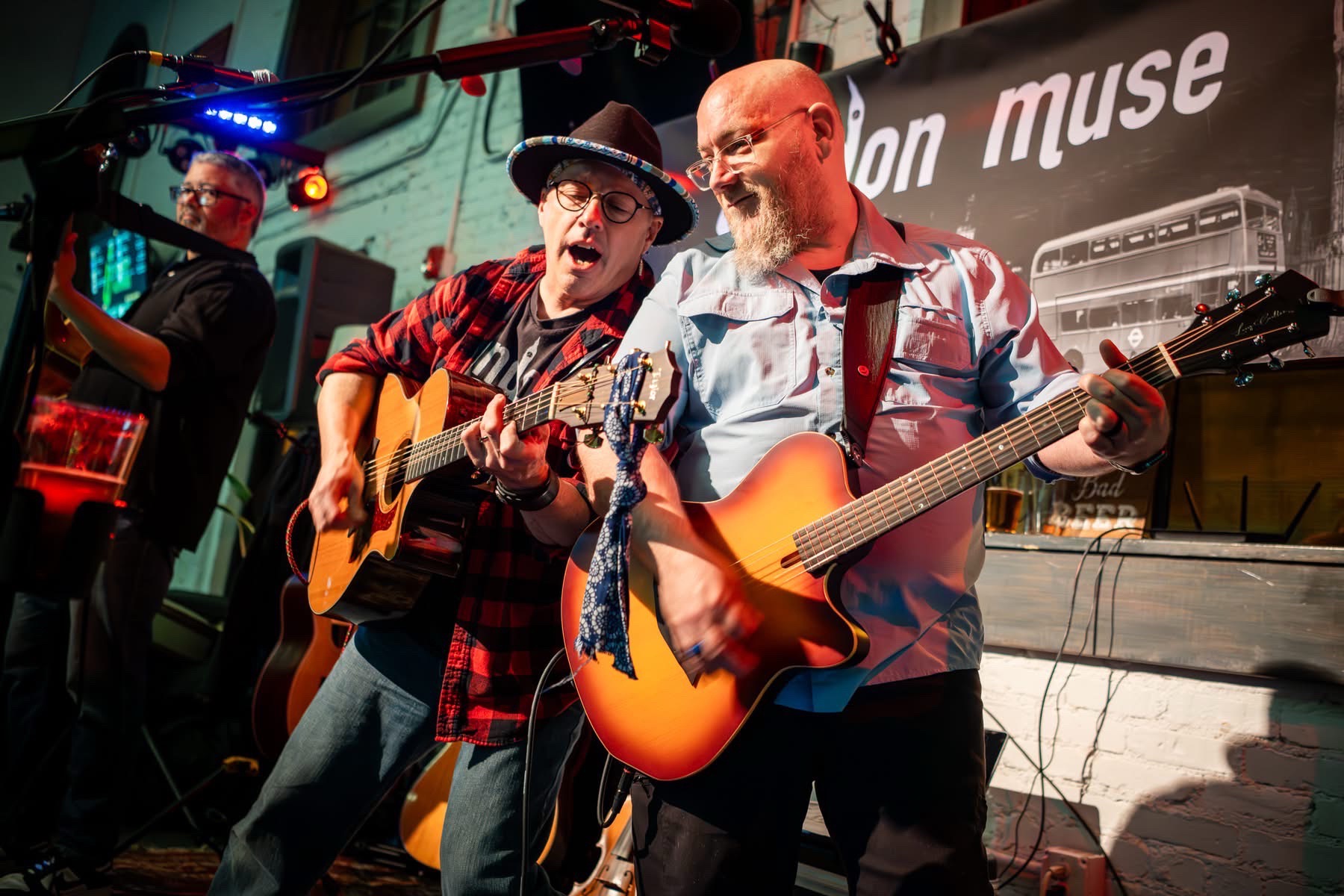 Two men playing guitars on stage, one singing, with a band in the background and a banner reading 'Muse'.