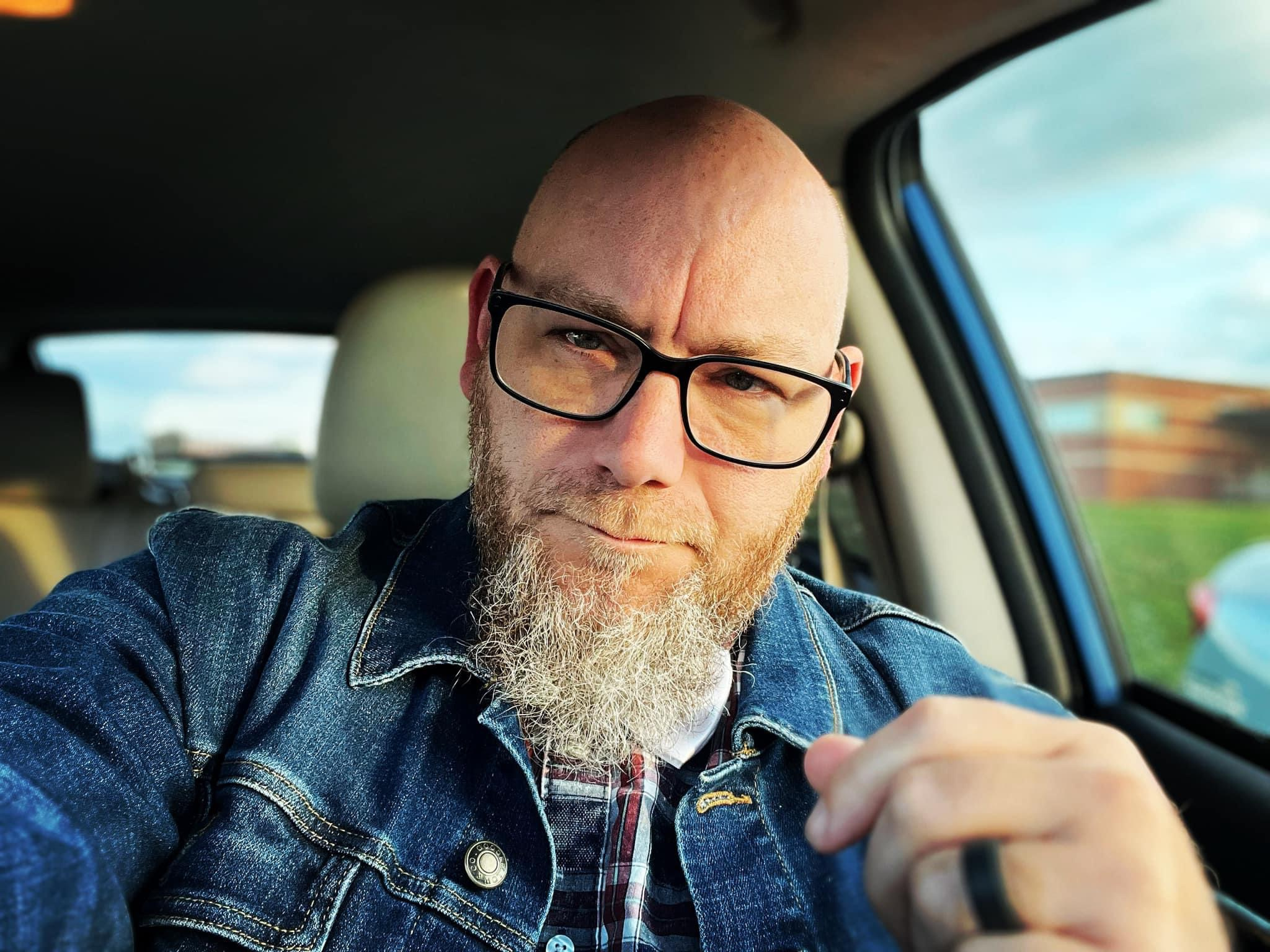 Man with glasses and a beard sitting in a car, looking at the camera, with a serious expression.
