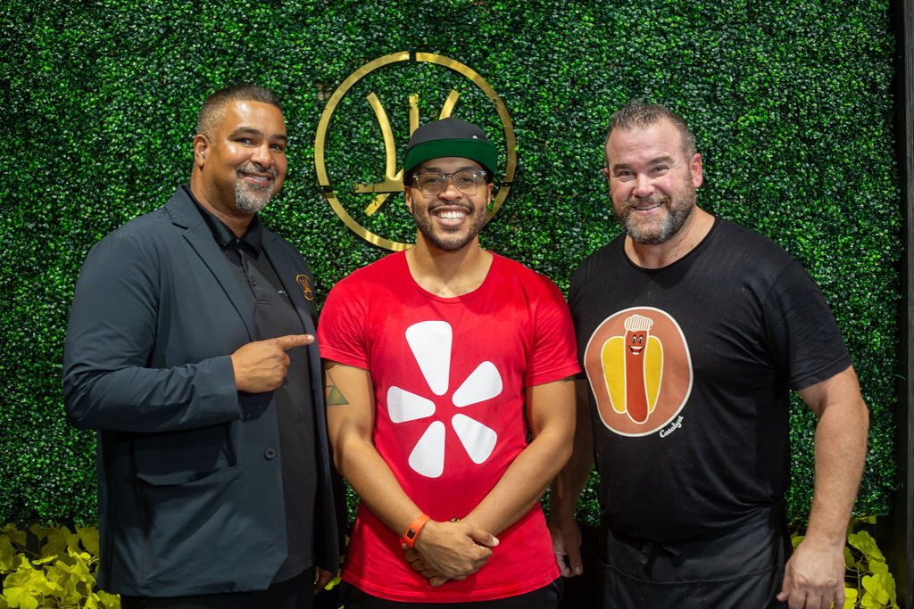 Three men standing in front of a green wall with a logo, smiling, one wearing a red shirt, others in dark clothing.