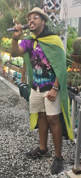Person smiling, wearing a hat, colorful shirt, shorts, and sandals, holding a microphone outdoors with plants and a sign nearby.