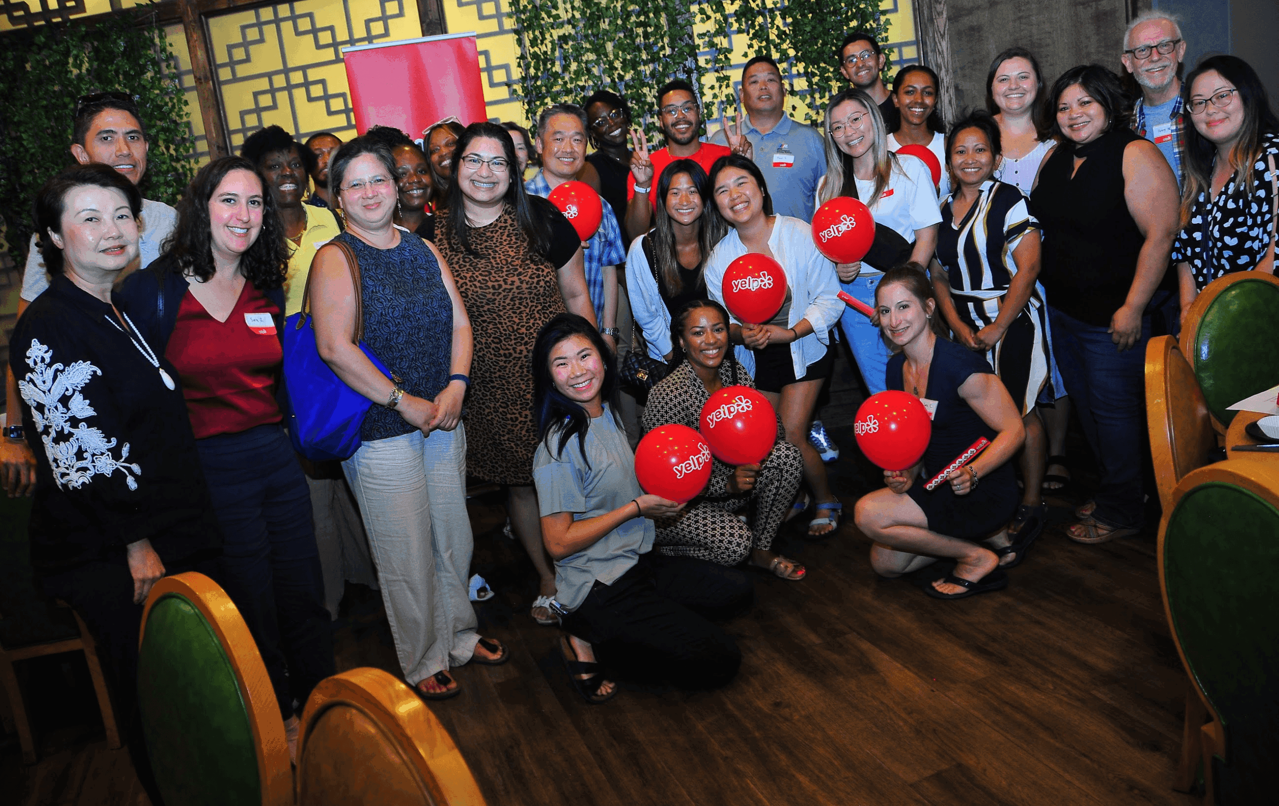 Group of people smiling indoors, holding red balloons, with chairs and decorative background, in a celebration setting.
