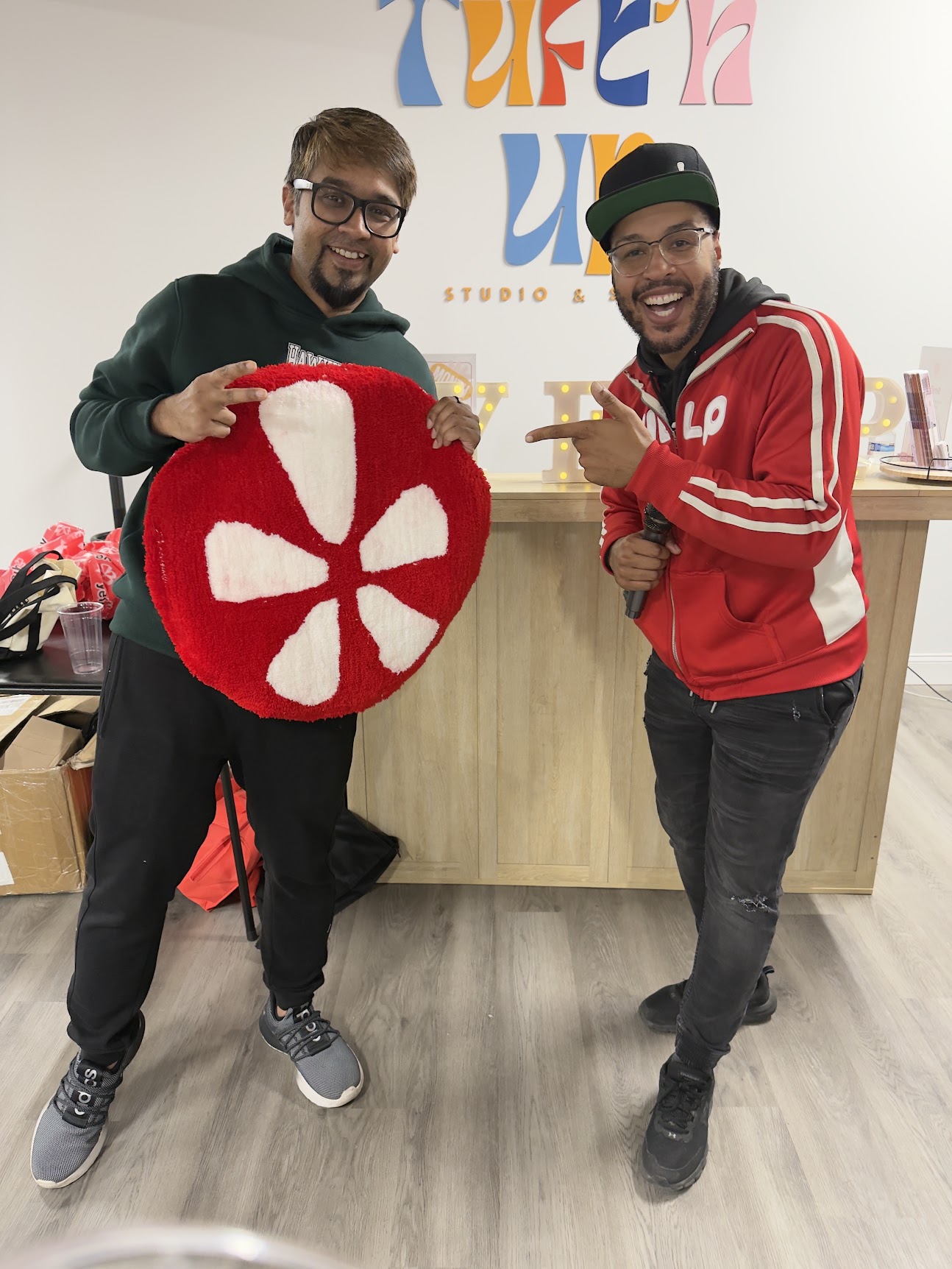 Two men smiling indoors, one holding a red and white circular object, the other pointing at it, with colorful wall art behind them.