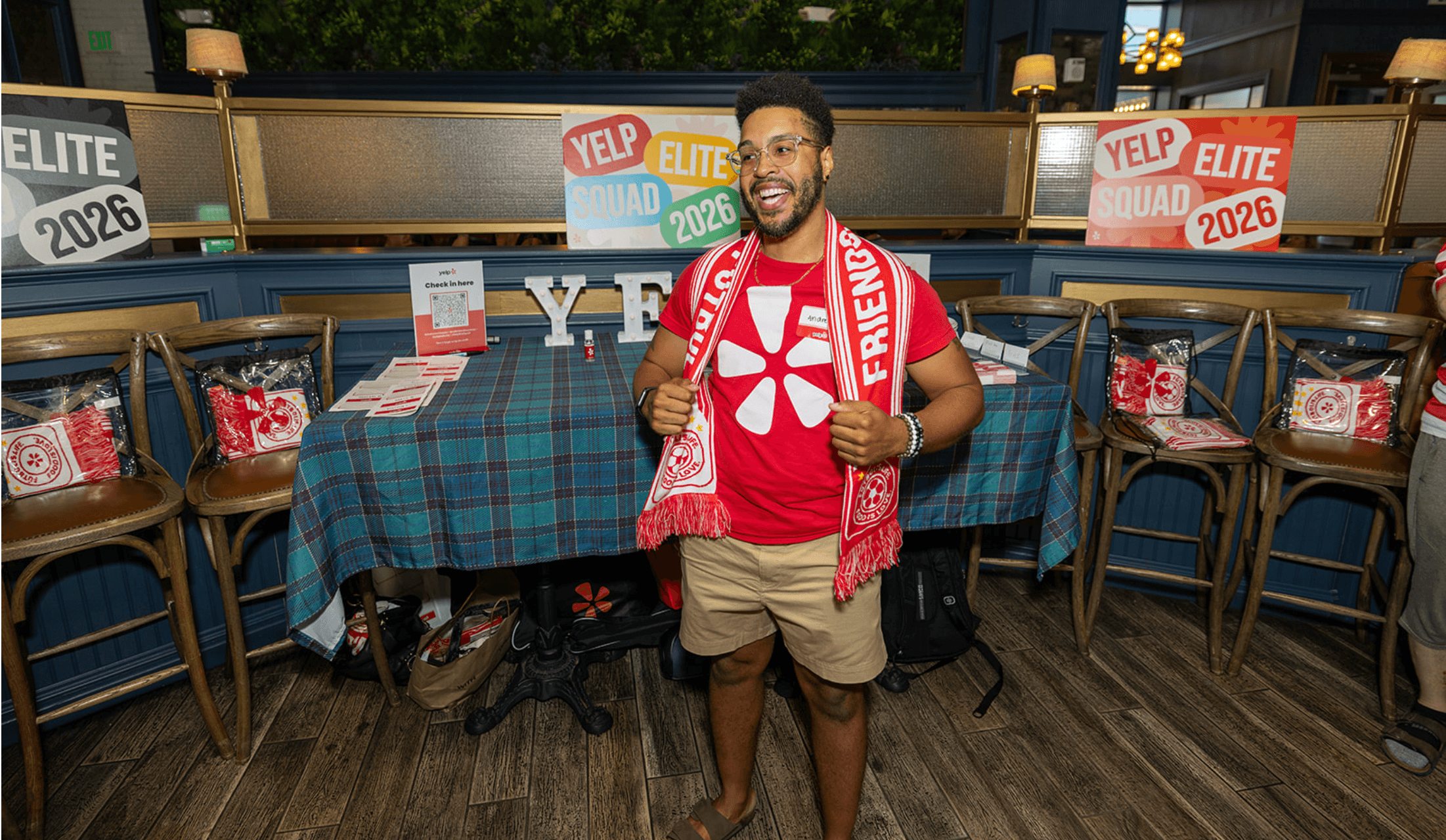 Man smiling at campaign event, wearing a red shirt and holding a water bottle, standing in front of campaign signs and tables.