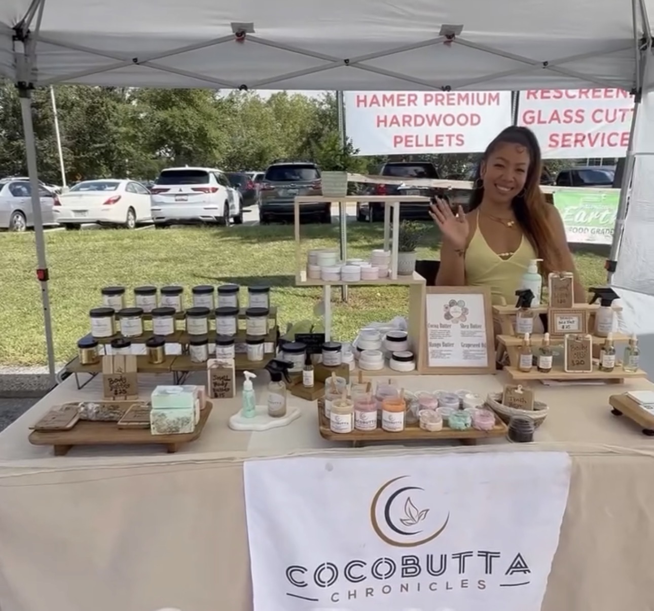 Woman smiling at a booth with jars and products, outdoor setting with cars and trees in background, booth banner reads Cocobutta Chronicles.
