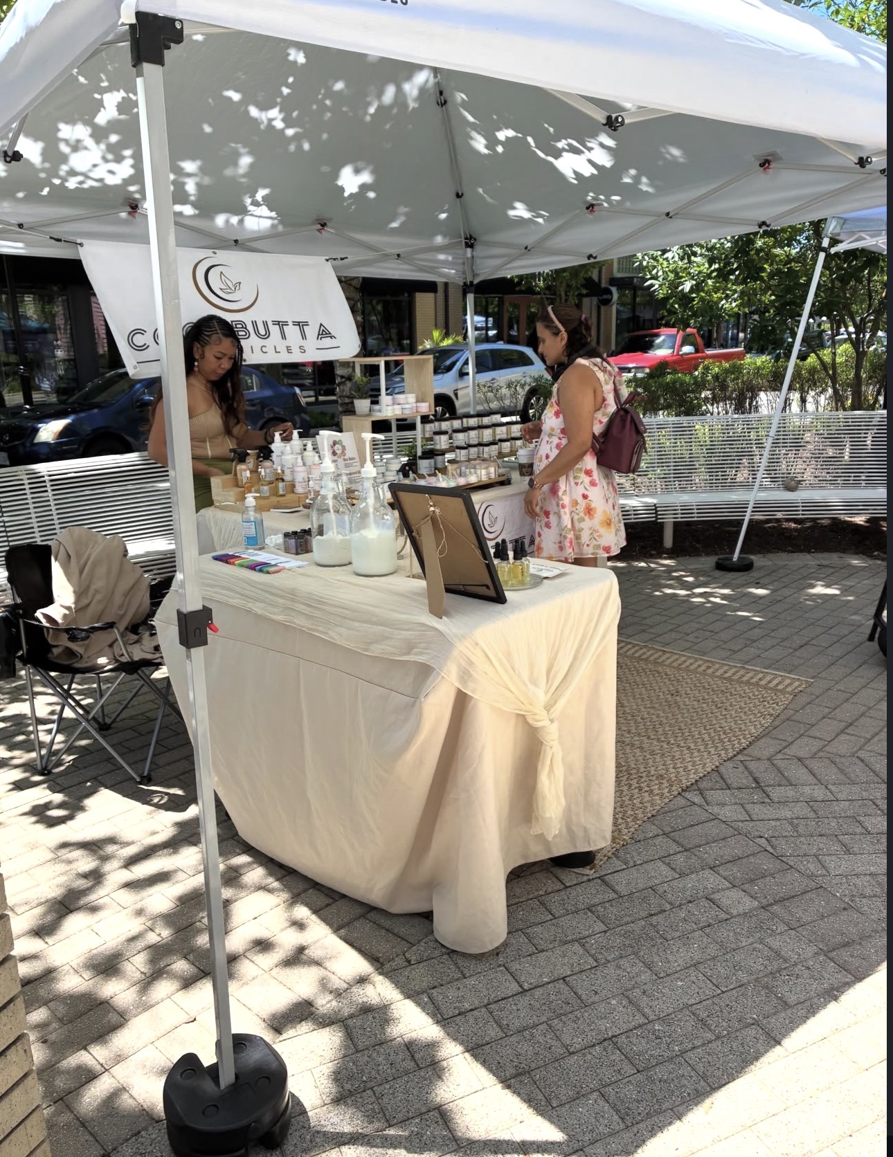 Outdoor market stall with a table covered in a white cloth, displaying bottles and items under a canopy, with two women nearby.