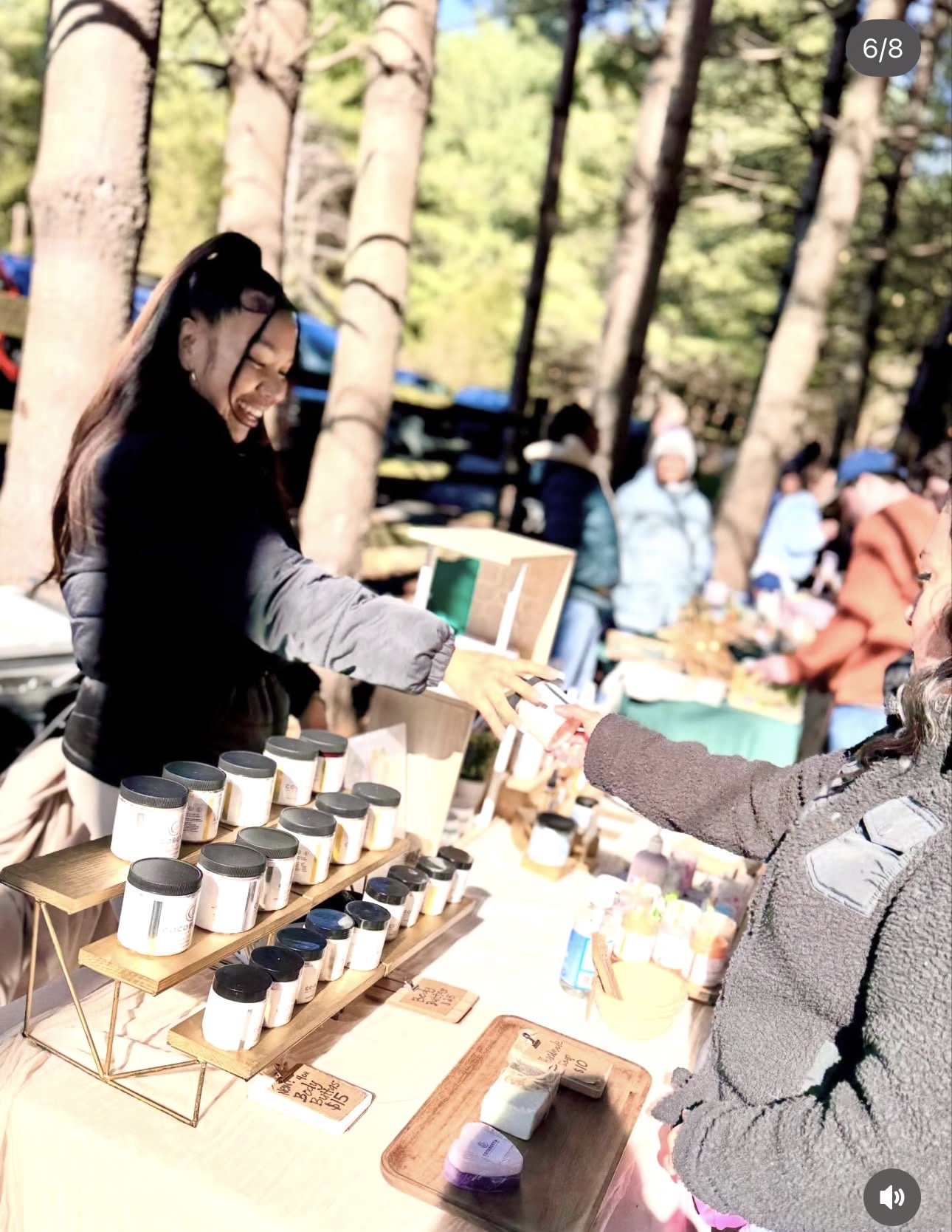Woman smiling at outdoor market stall with cups and products, trees and people in background, sunny day.