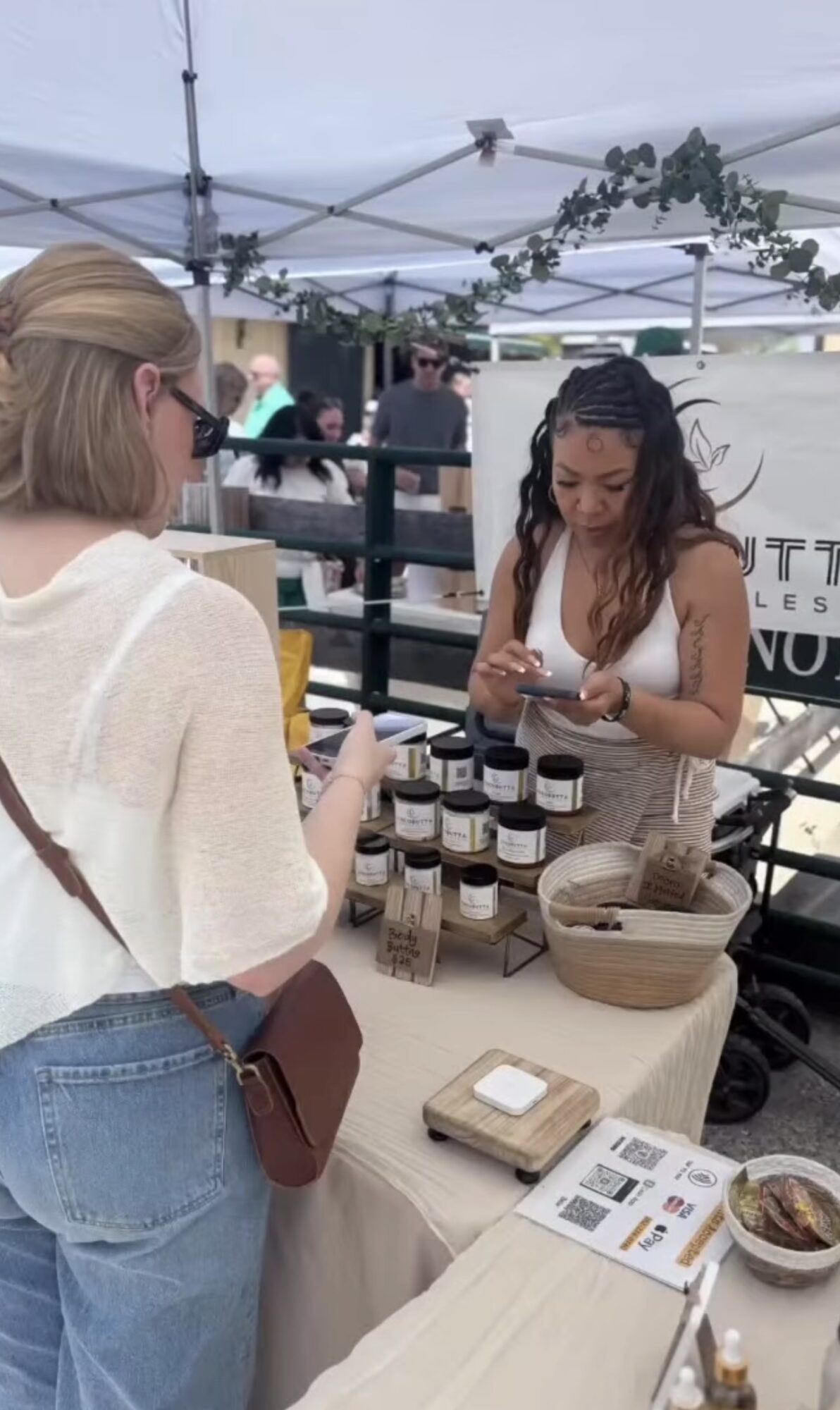 Two women at an outdoor market stall with jars and a bowl, one using a smartphone, under a canopy.