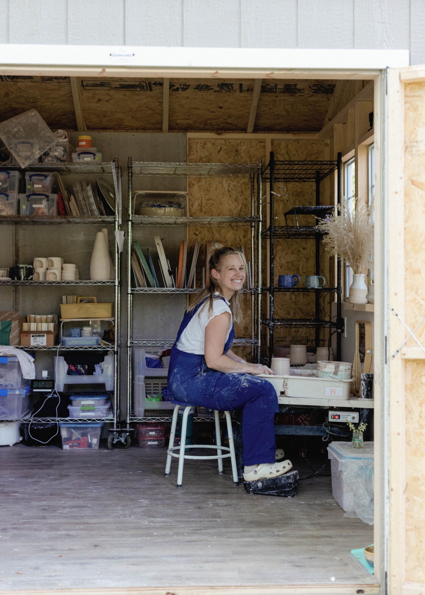 Young woman sitting on a stool in a workshop, surrounded by shelves with tools and supplies, smiling at the camera.