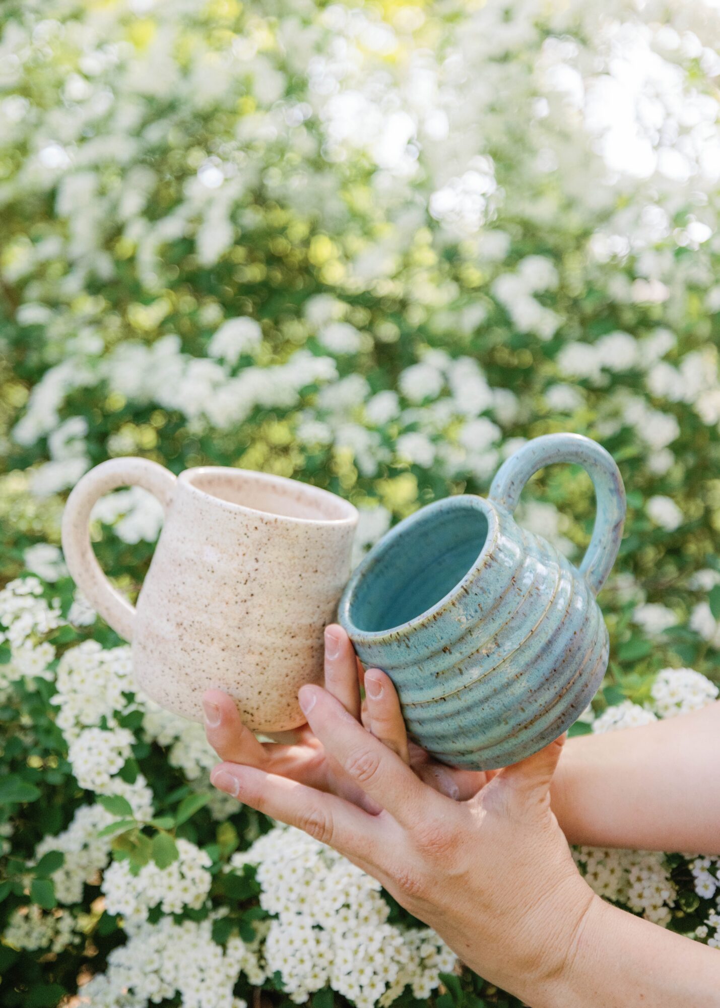 Person holding two ceramic mugs outdoors with white flowers and green foliage in background.