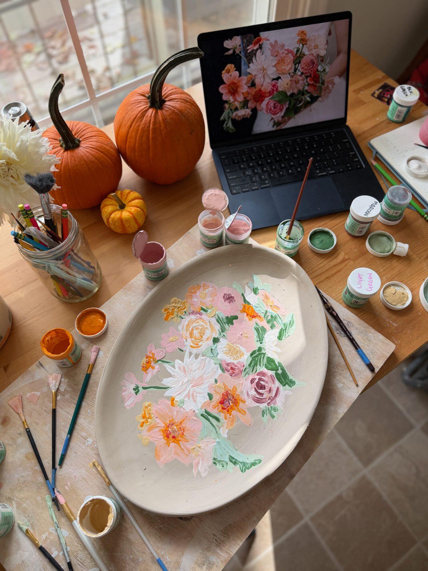 Painting of colorful flowers on a large oval ceramic plate, surrounded by paint jars, brushes, pumpkins, and a laptop with floral wallpaper.