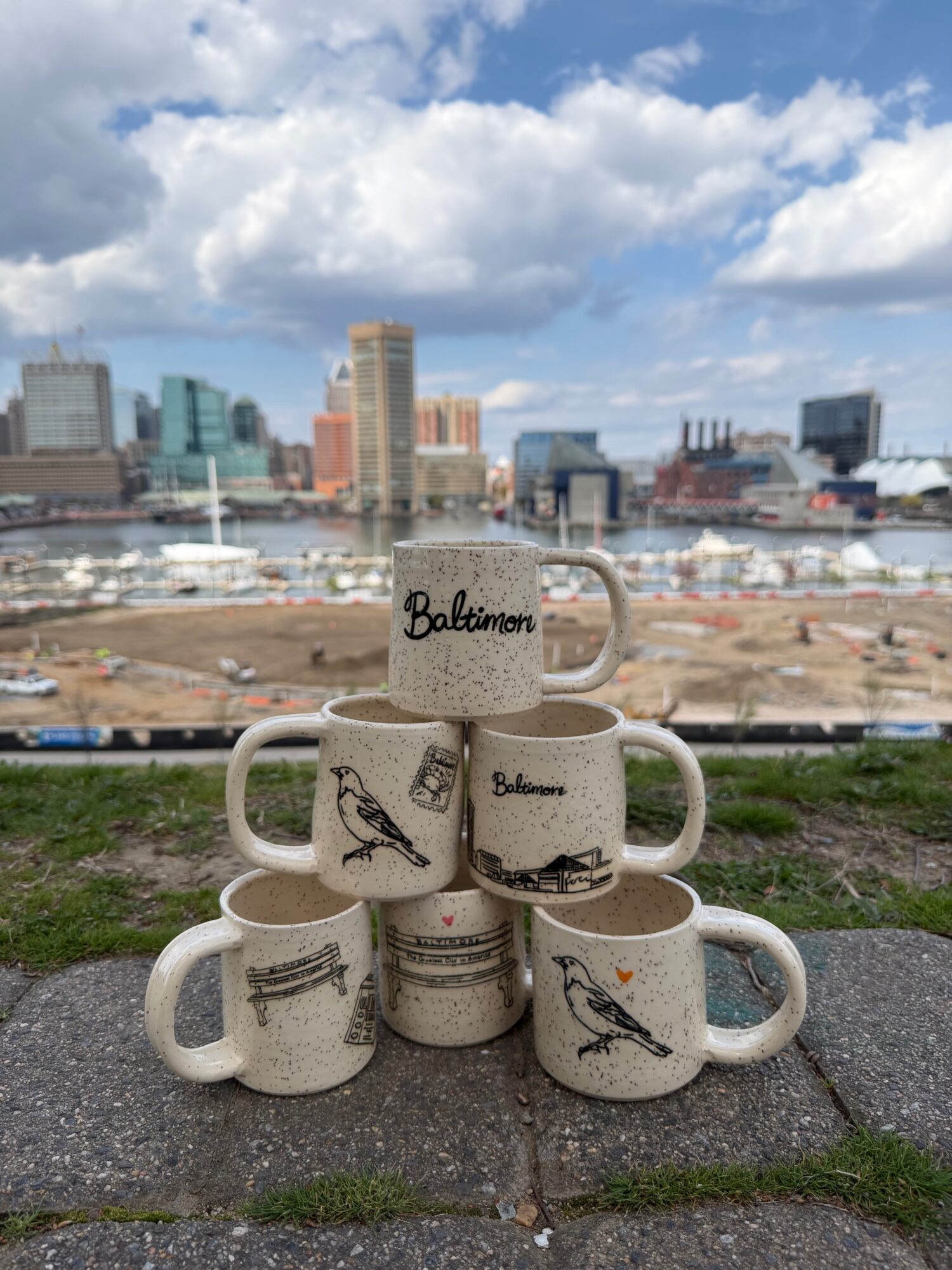 Six stacked coffee mugs with Baltimore-themed designs on a ledge, city skyline and sky in background.