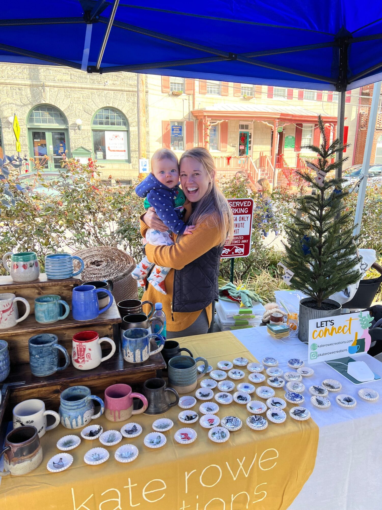 Two women stand behind a table with candles and a small Christmas tree, outdoors on a sunny day.