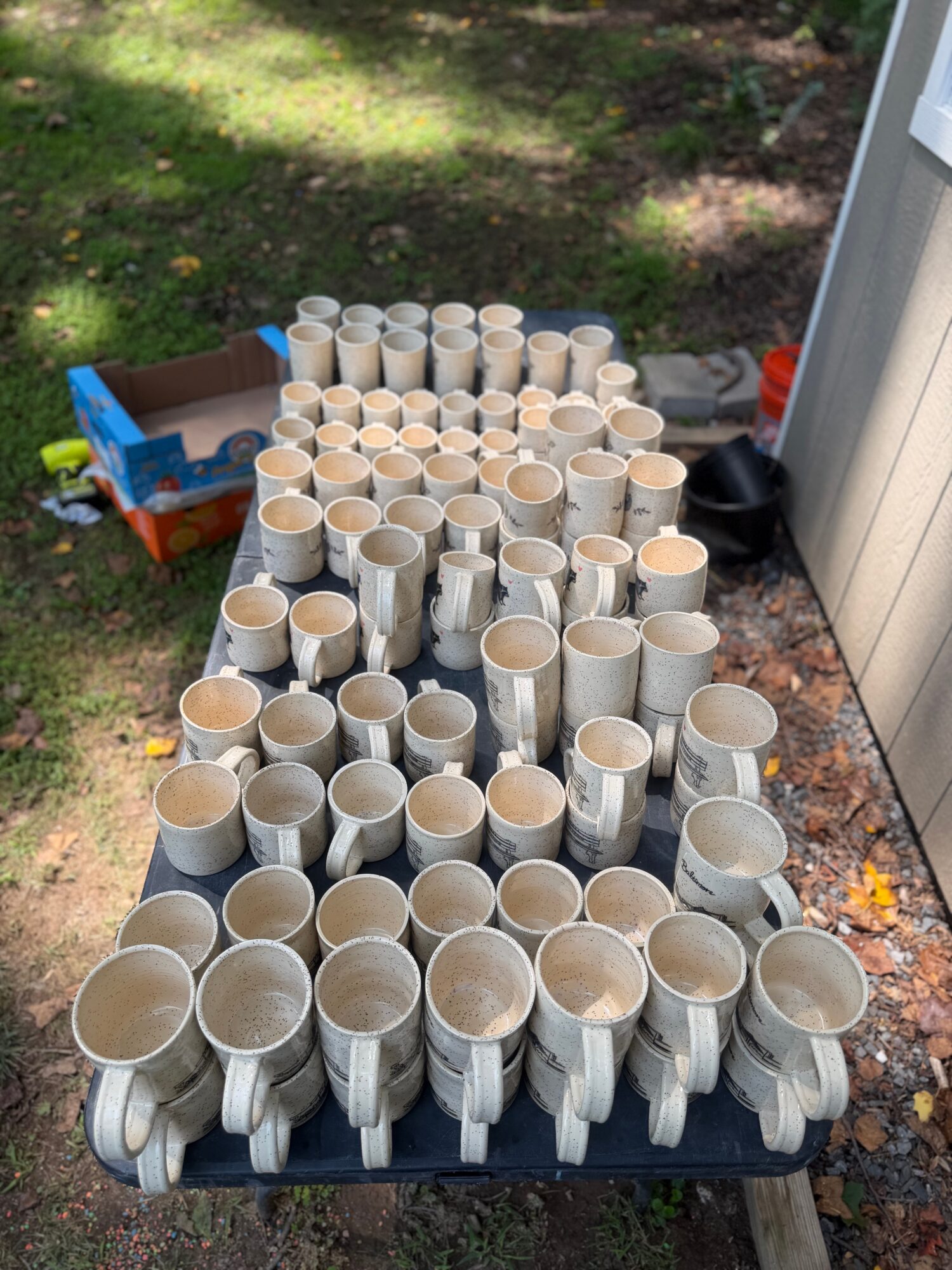 Multiple white ceramic mugs arranged on a black table outdoors, with a grassy area and a shed in the background.