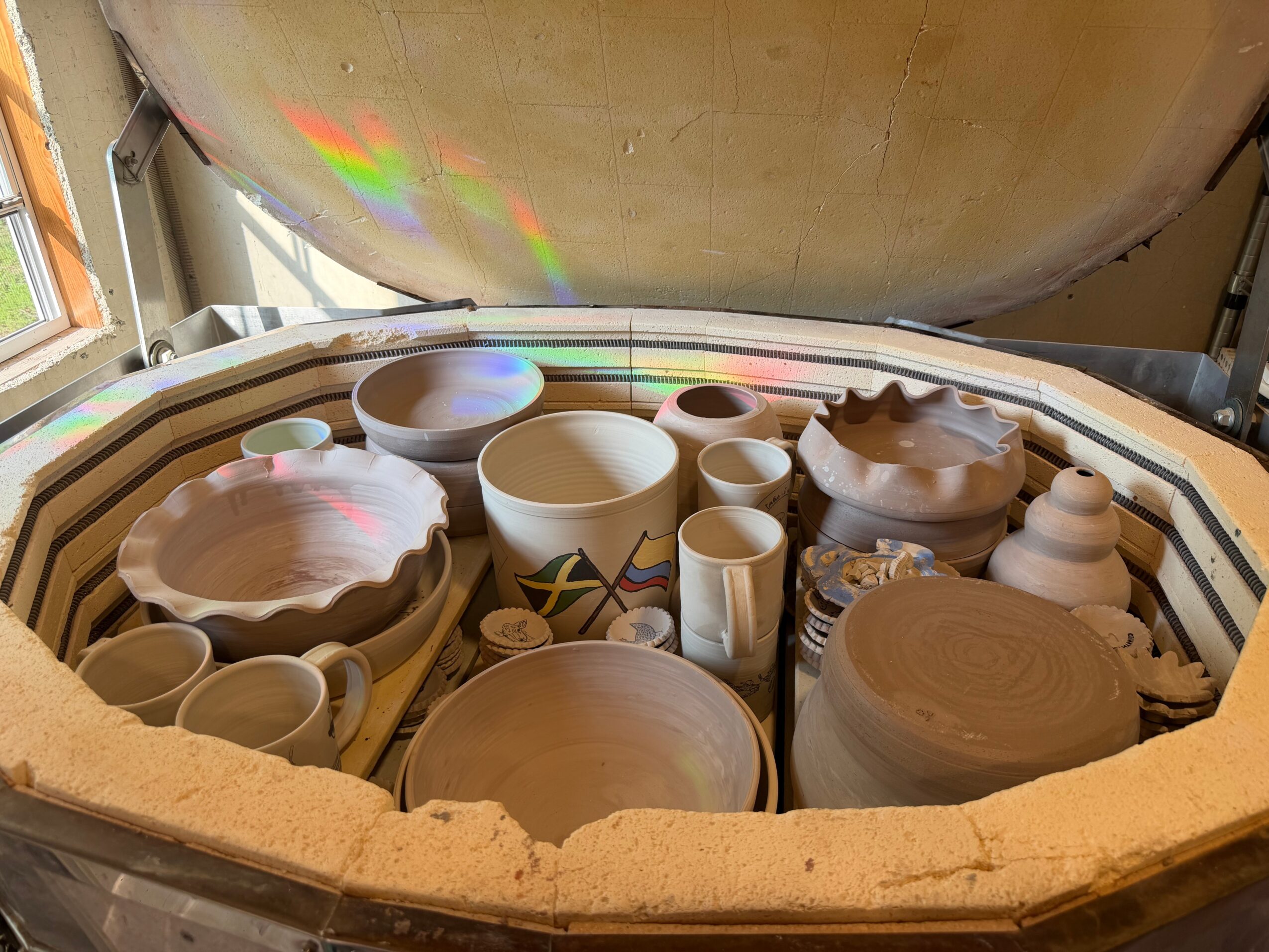 Open kiln with unglazed pottery including bowls, cups, and vases inside, with a rainbow reflection on the lid.