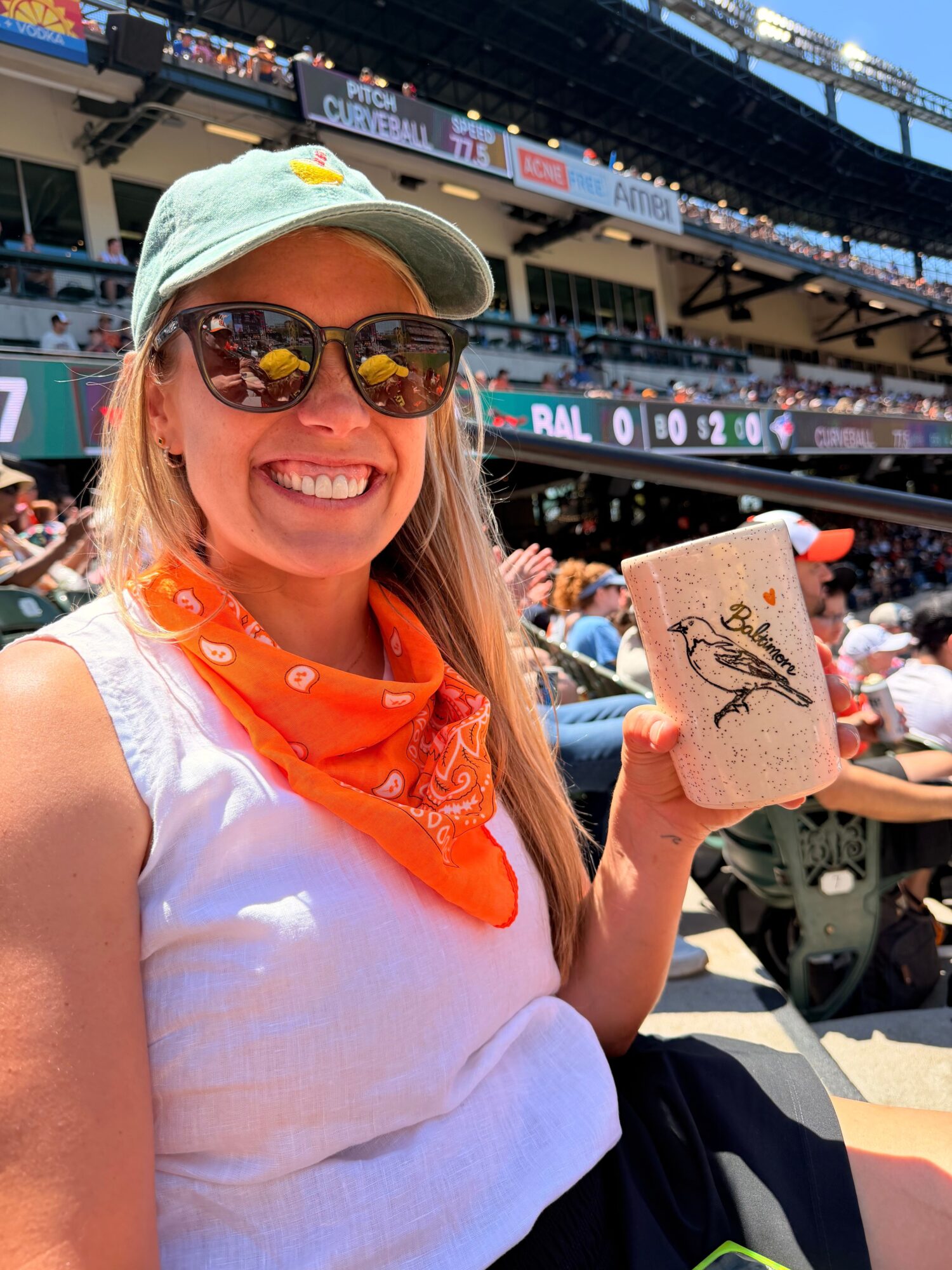 Woman smiling, wearing sunglasses and a cap, holding a pink phone case, at a sports event or stadium.
