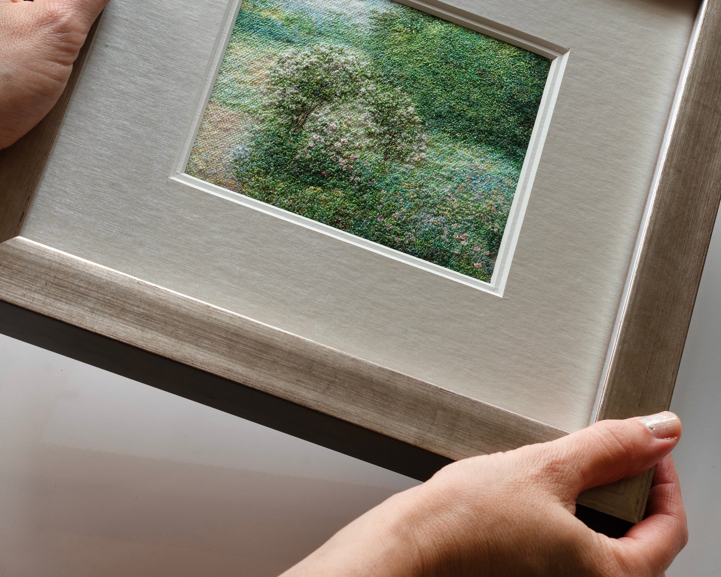 Person holding a framed watercolor painting of a green landscape, with hands visible at the bottom corners.