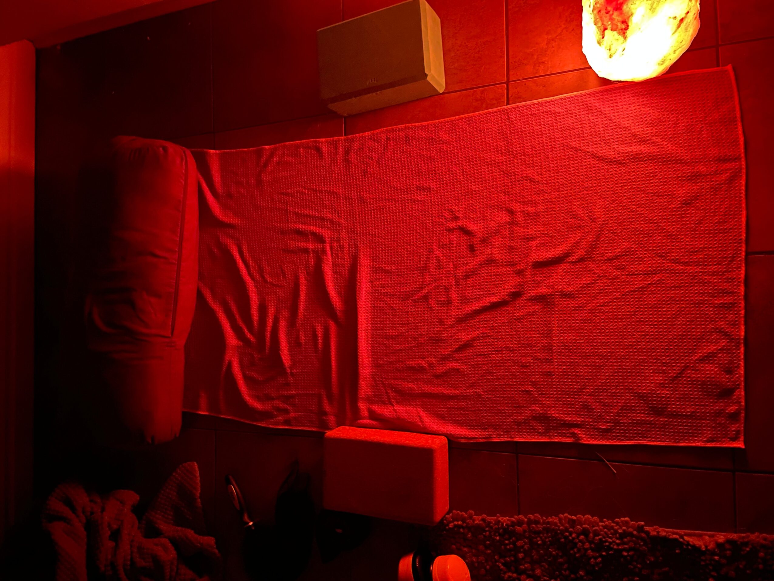 Red bed with a blanket, a pillow, and a glowing salt lamp on a dark floor, viewed from the side.