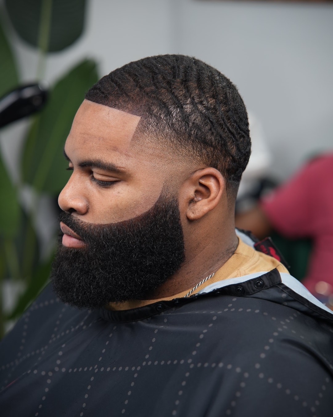 Side profile of a man with a textured fade haircut and full beard, looking downward, in a salon setting.