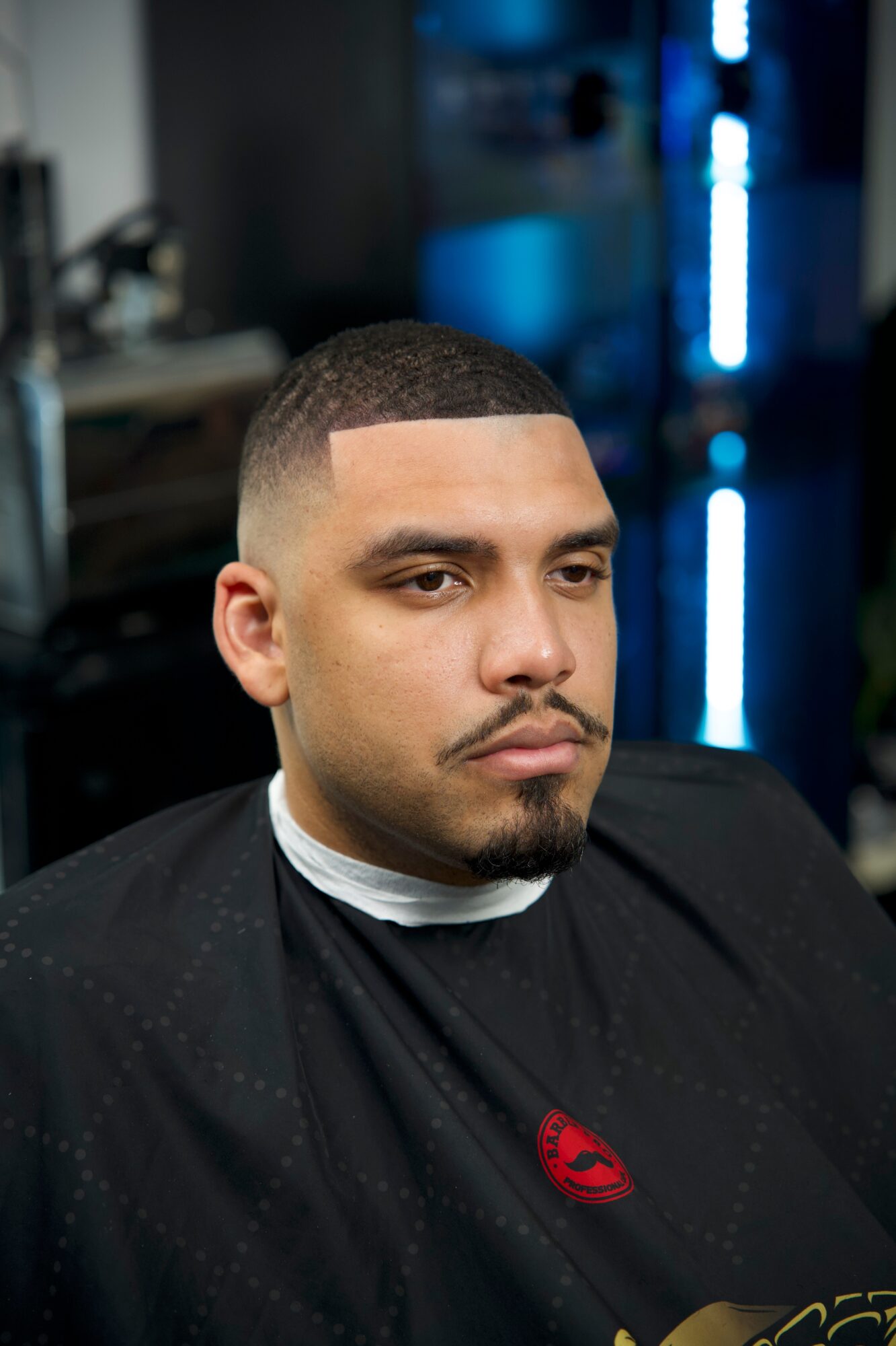 Young man with short hair and a goatee, wearing a black barber cape, sitting in a barbershop, looking serious.