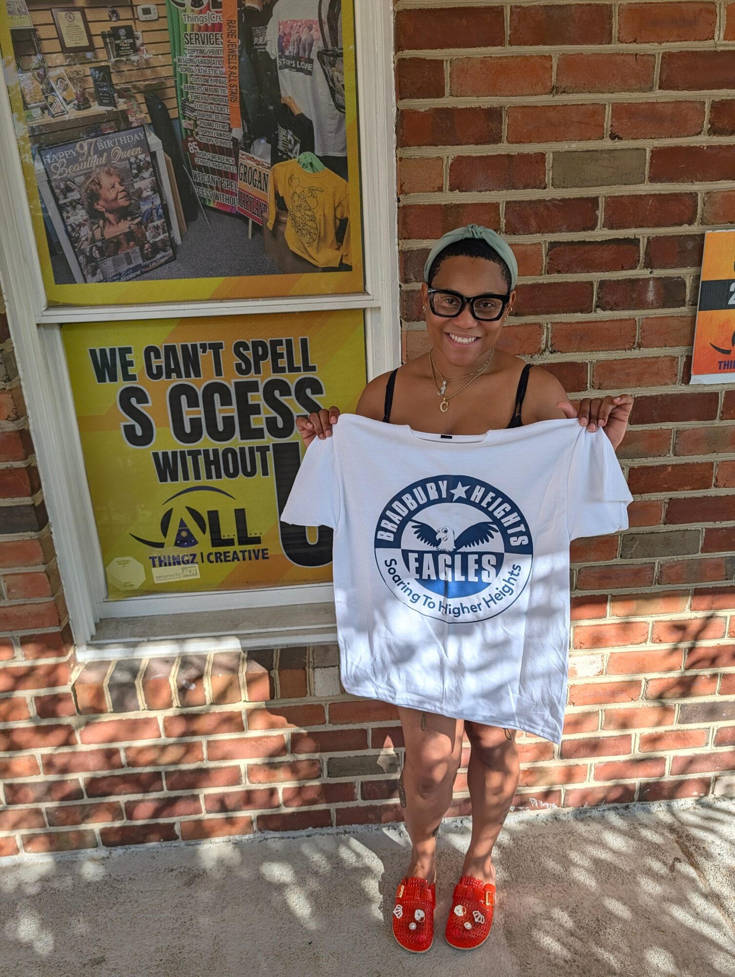 Person with short hair and glasses holding a white T-shirt with a logo, standing outside a brick building.