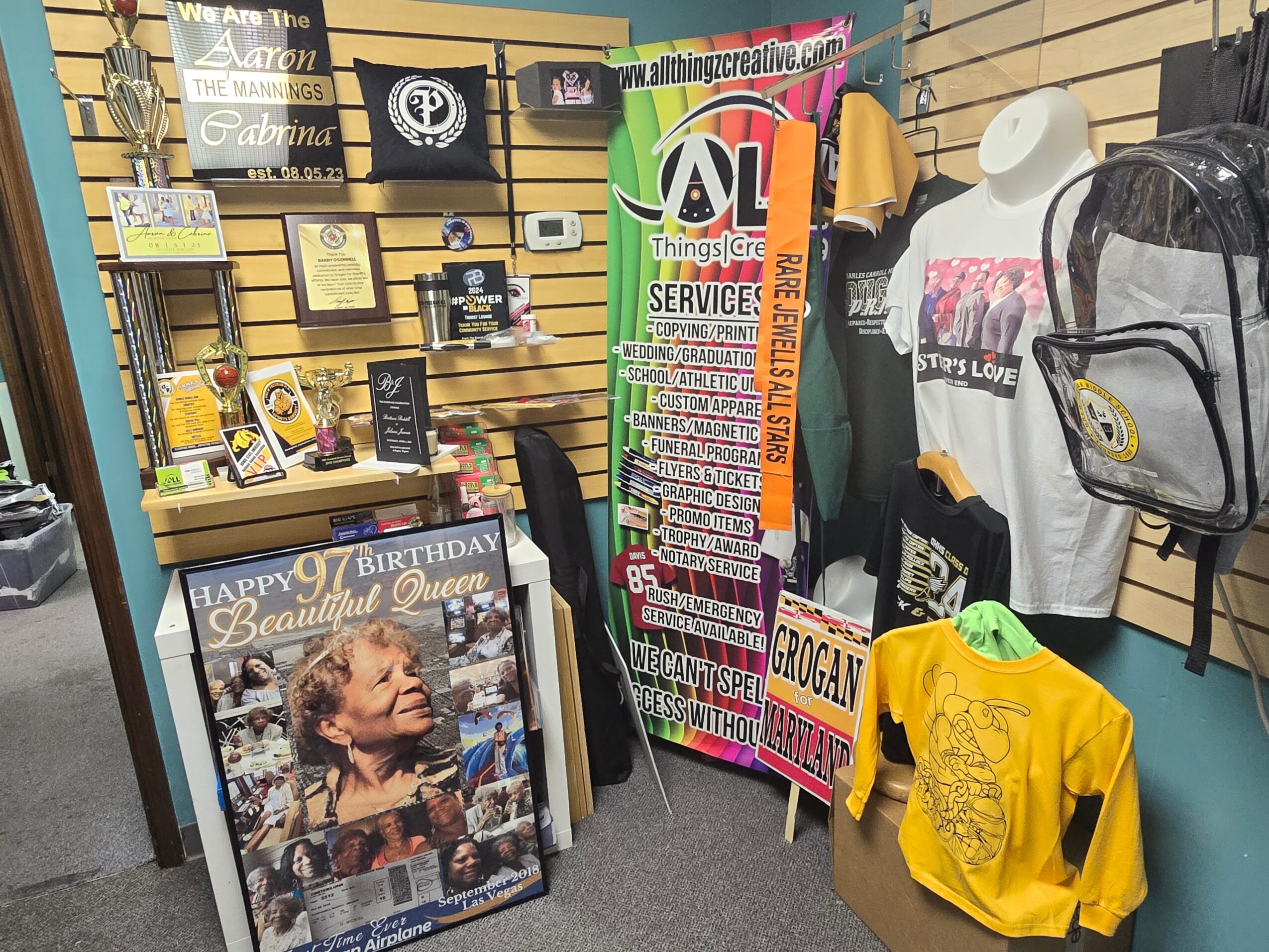 Display of framed photos, clothing, and signs in a store, including a birthday poster and a colorful service sign.