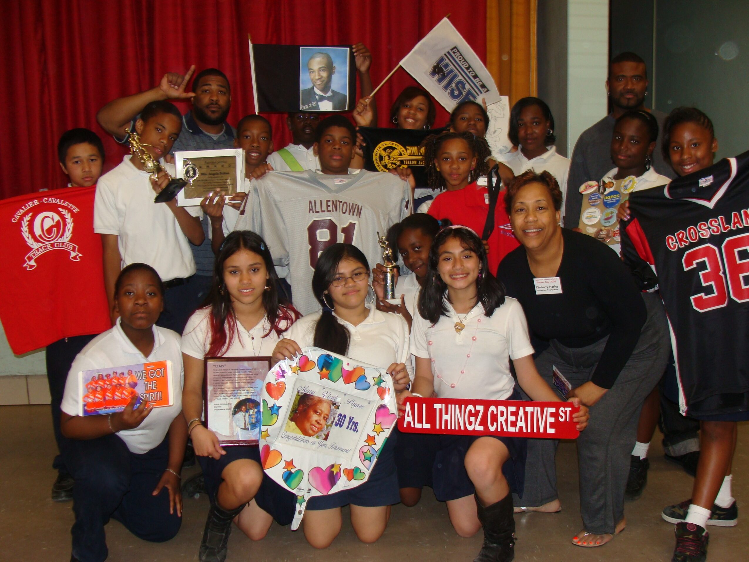 Group of children and adults holding signs and artwork in a room with red curtains, some smiling and posing for the photo.