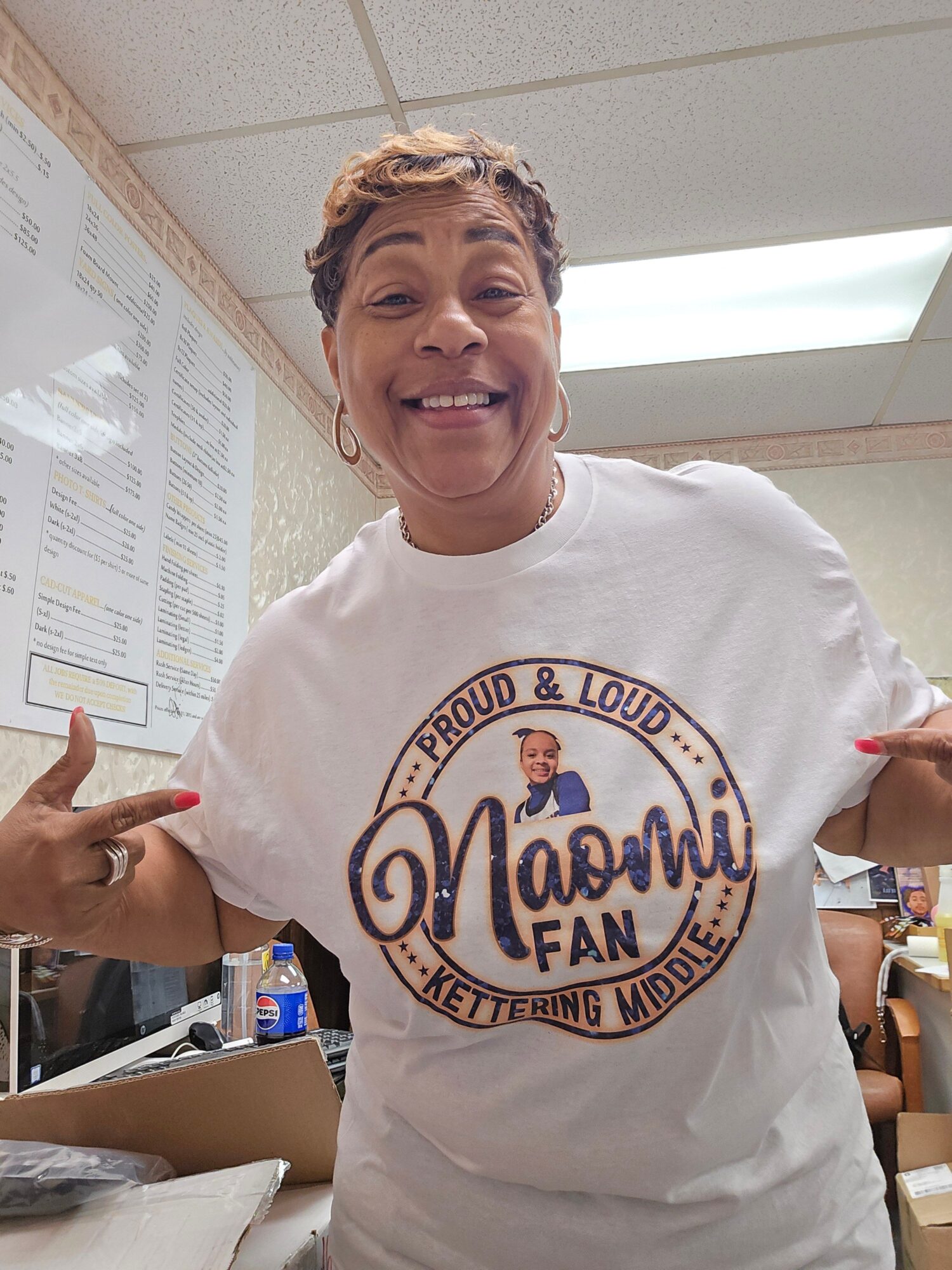 Person smiling and pointing at a white T-shirt with a logo, in an indoor setting with a ceiling light and window.