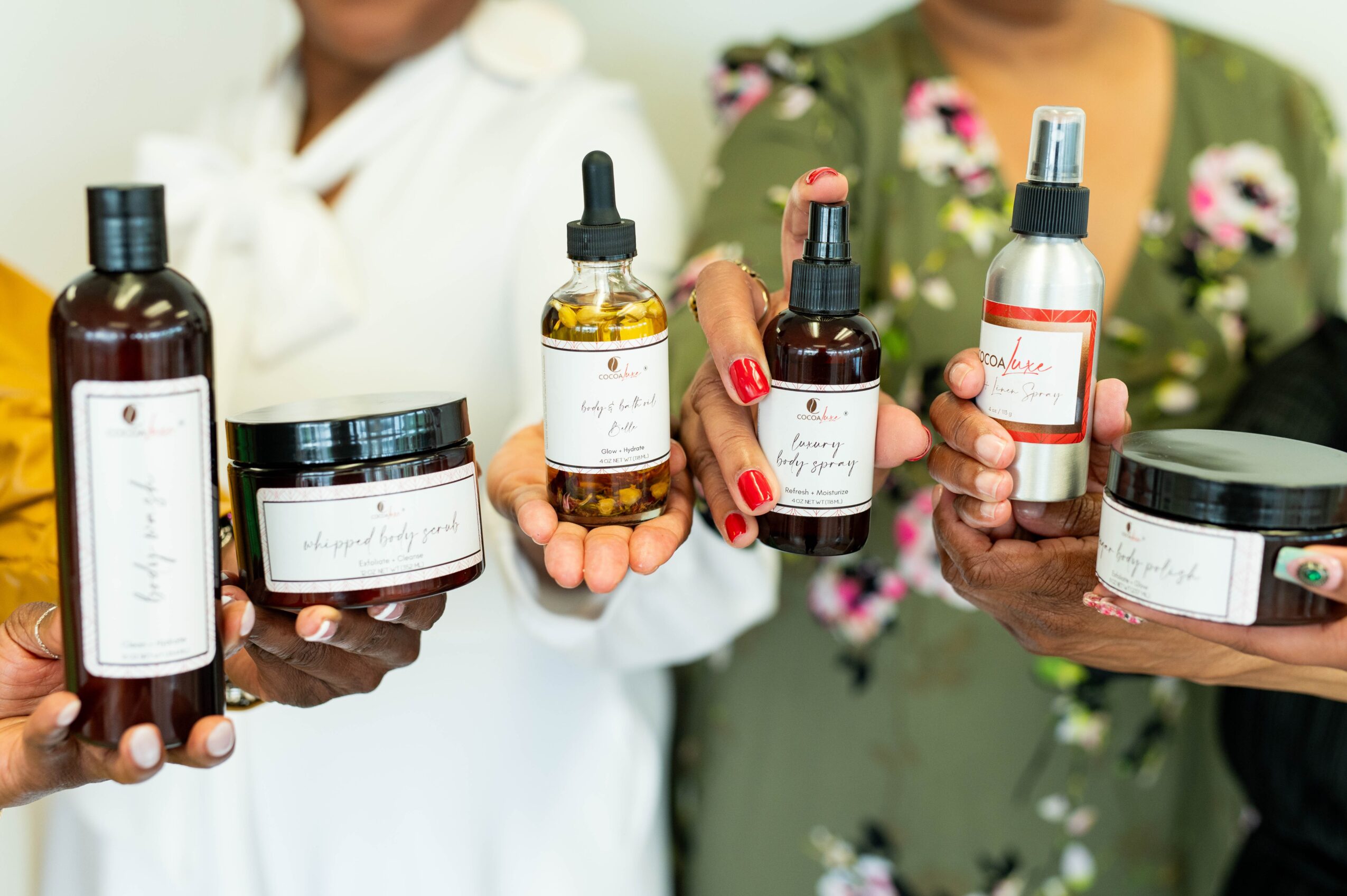 People holding skincare products, including bottles and jars, with blurred background of women in floral and white clothing.