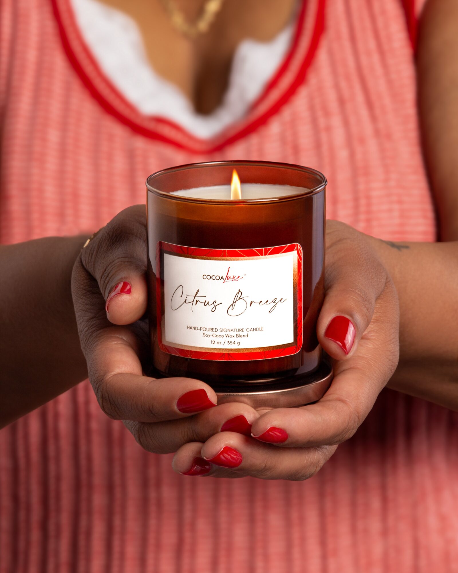 Person holding a lit candle in a glass jar with a label, wearing a pink top with white lace, red nail polish.