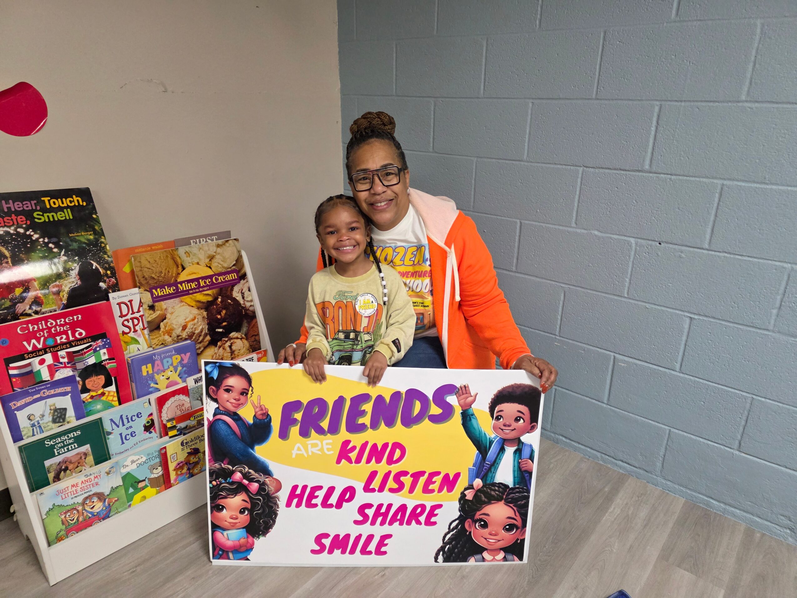 Two girls holding a colorful sign with cartoon characters, smiling indoors near a bookshelf, with a blue wall behind them.