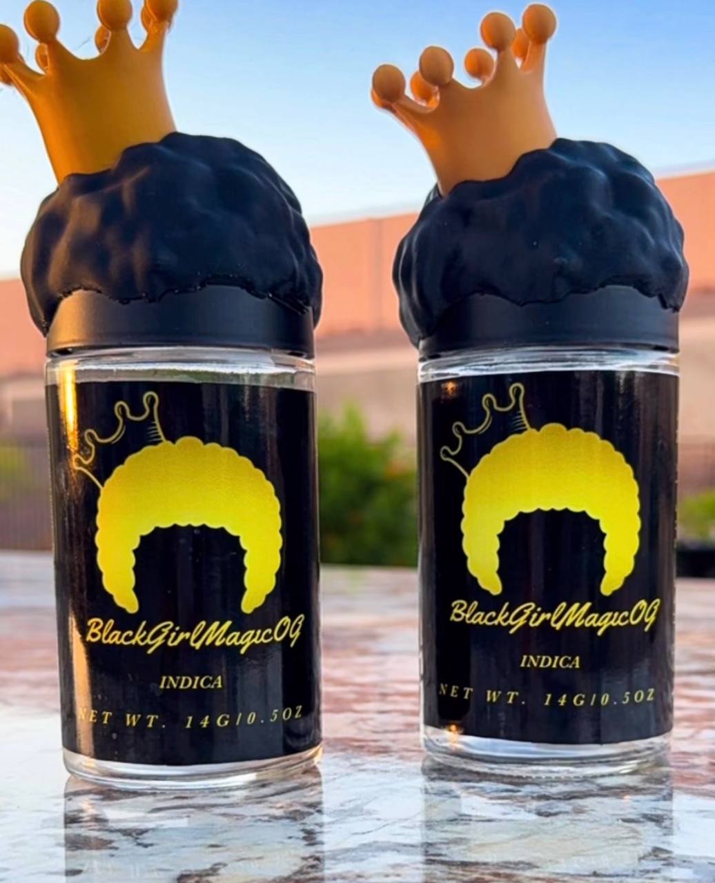 Two bottles with black labels and yellow hair illustrations, topped with crown-shaped caps, outdoors on a table.