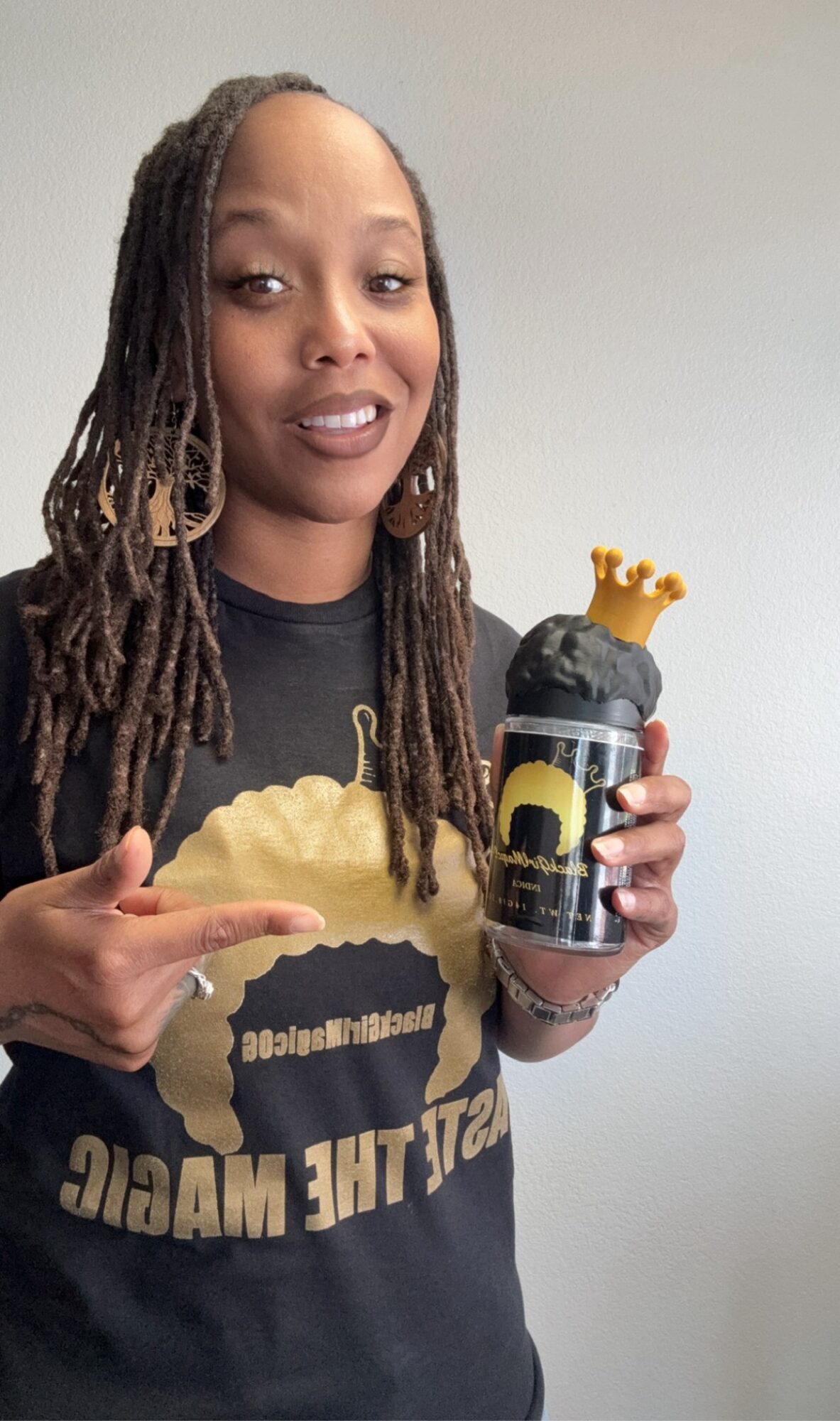 Person with long dreadlocks holding a bottle with a crown cap, wearing a black T-shirt with a graphic and text, pointing at the bottle.