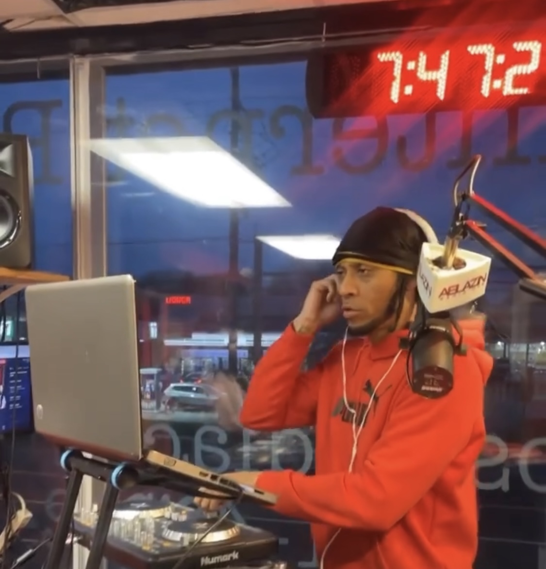 Person wearing a black cap and red hoodie, sitting in a radio station, with a computer and microphone, looking at the screen.