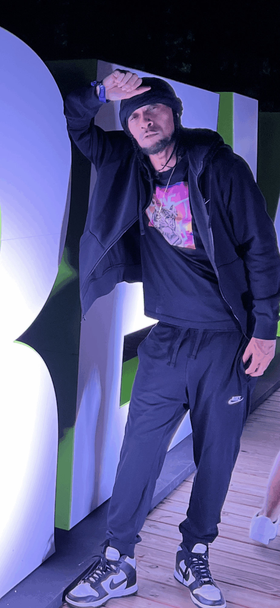 Person with dark hair and beard wearing a black jacket, black pants, and sneakers, standing indoors near colorful panels.