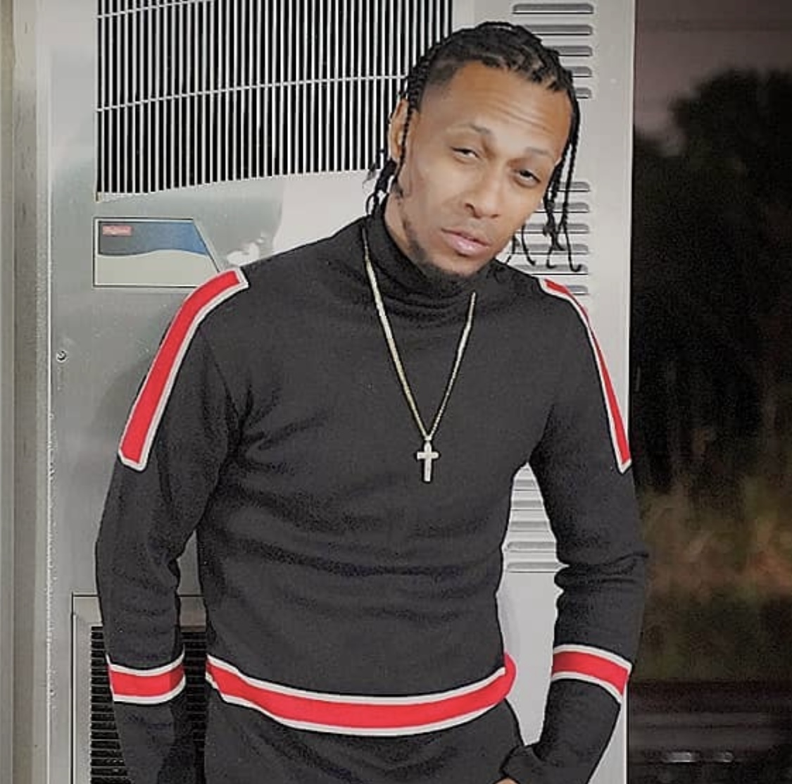 Young man with braided hair wearing a black sweater with red and white stripes, standing outdoors near a metal structure.