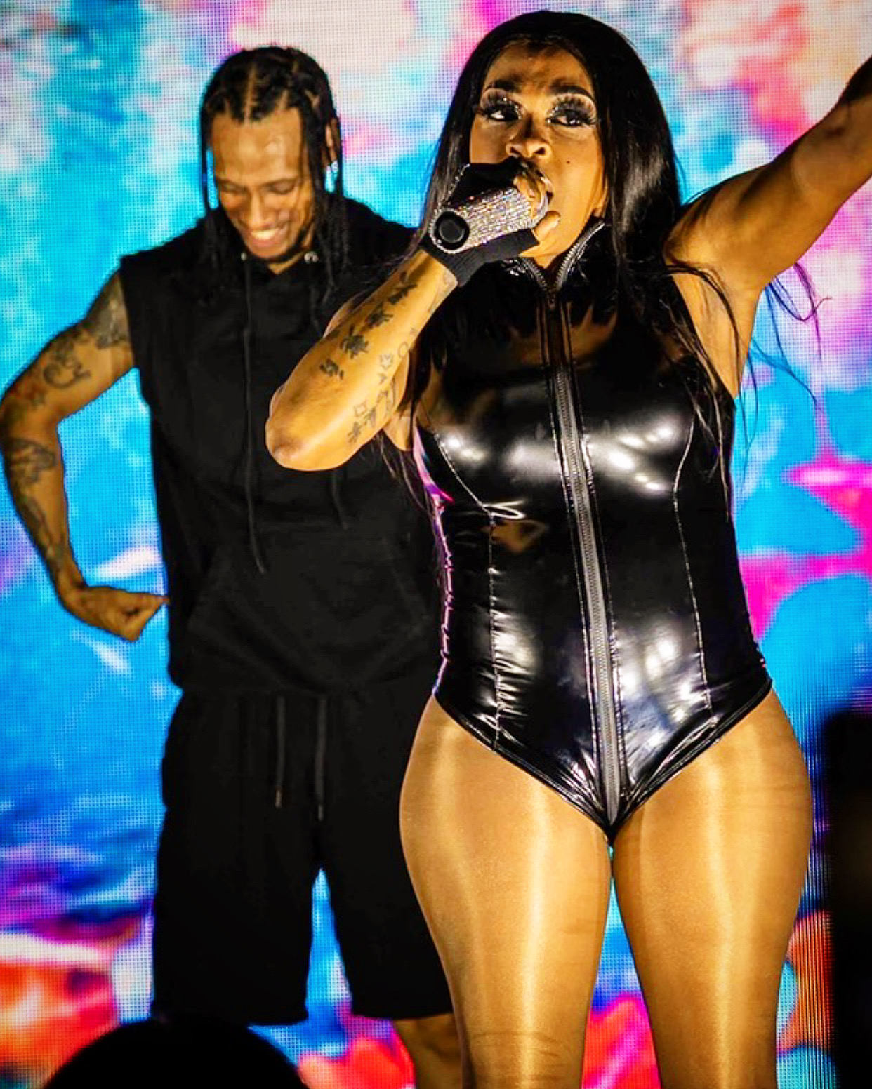 Performer in a shiny black outfit singing into a microphone, with a person in black behind her, colorful background.